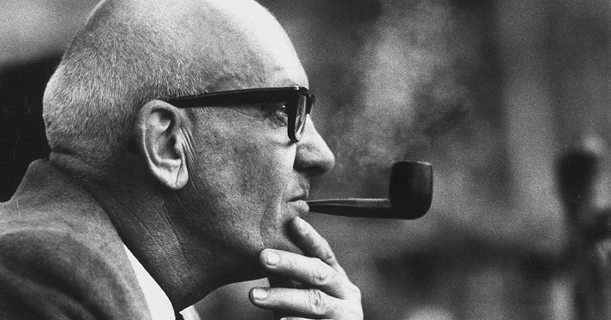 A profile of Mr. Kauffman wearing thick-rimmed glasses and smoking a pipe.