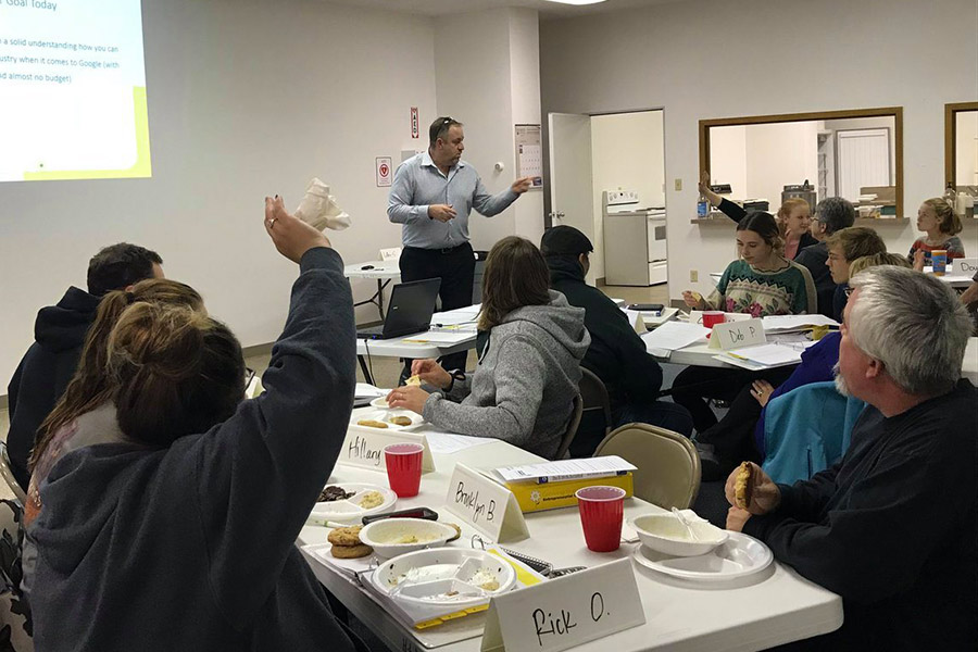 A facilitator stands at the front of a classroom full of participants and points to someone raising their hand.