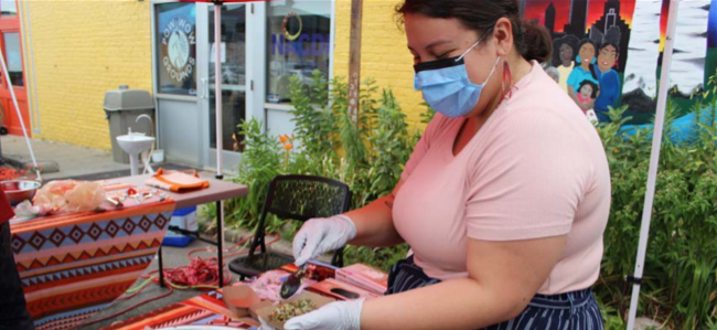 A person wearing a mask and gloves scoops food into a to-go container at an outdoor farmers market