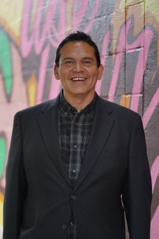 A photo of John Williams, smiling and standing in front of a graffitied wall, wearing a black jacket and dark blue patterned shirt.