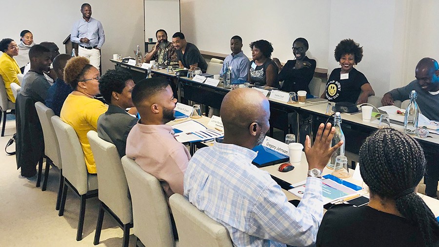 Nineteen people sit together in conversation at two long conference room tables at a Builders and Benefactors convening