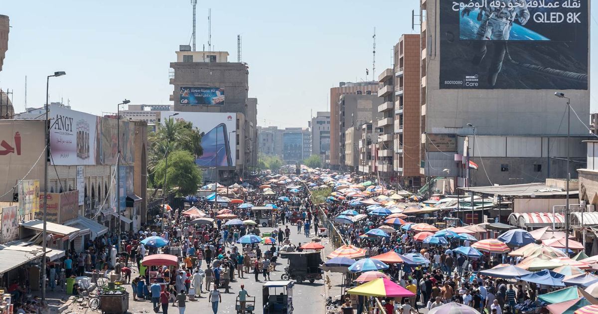 A wide-angle photo of Baghdad, Iraq, shows dozens of people walking down a city street holding umbrellas to block the sun. A building in the background features a large banner image of an astronaut.