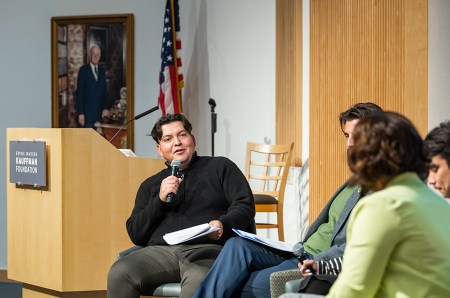 Michale Carmona moderates a panel while sitting on stage. A photo of Kauffman Foundation founder Ewing Marion Kauffman hangs on the wall behind him next to an American flag.