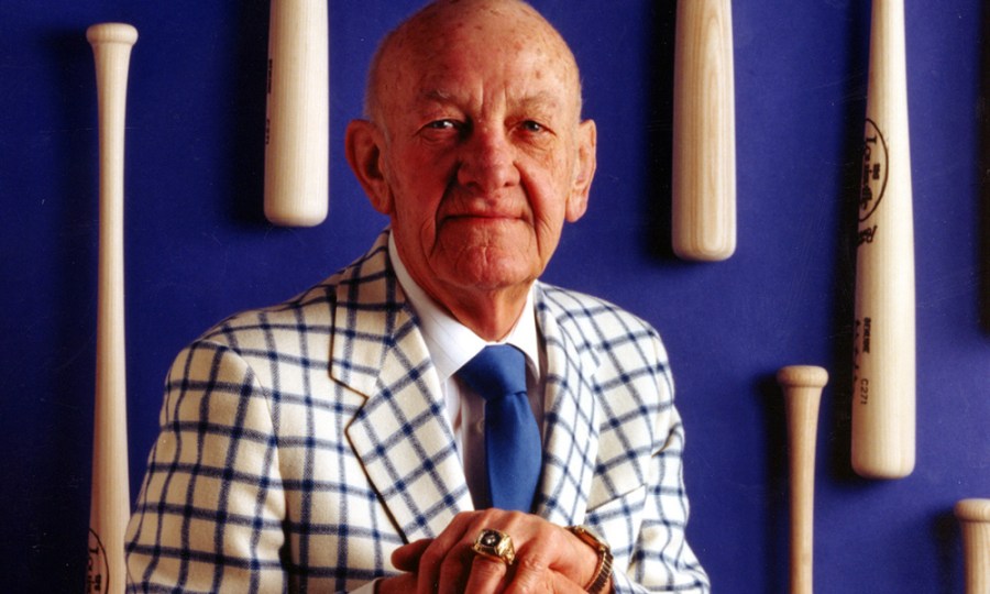 Ewing Kauffman sits in front of a royal blue wall with baseball bats attached to it. He wears a white and blue checkered suit.