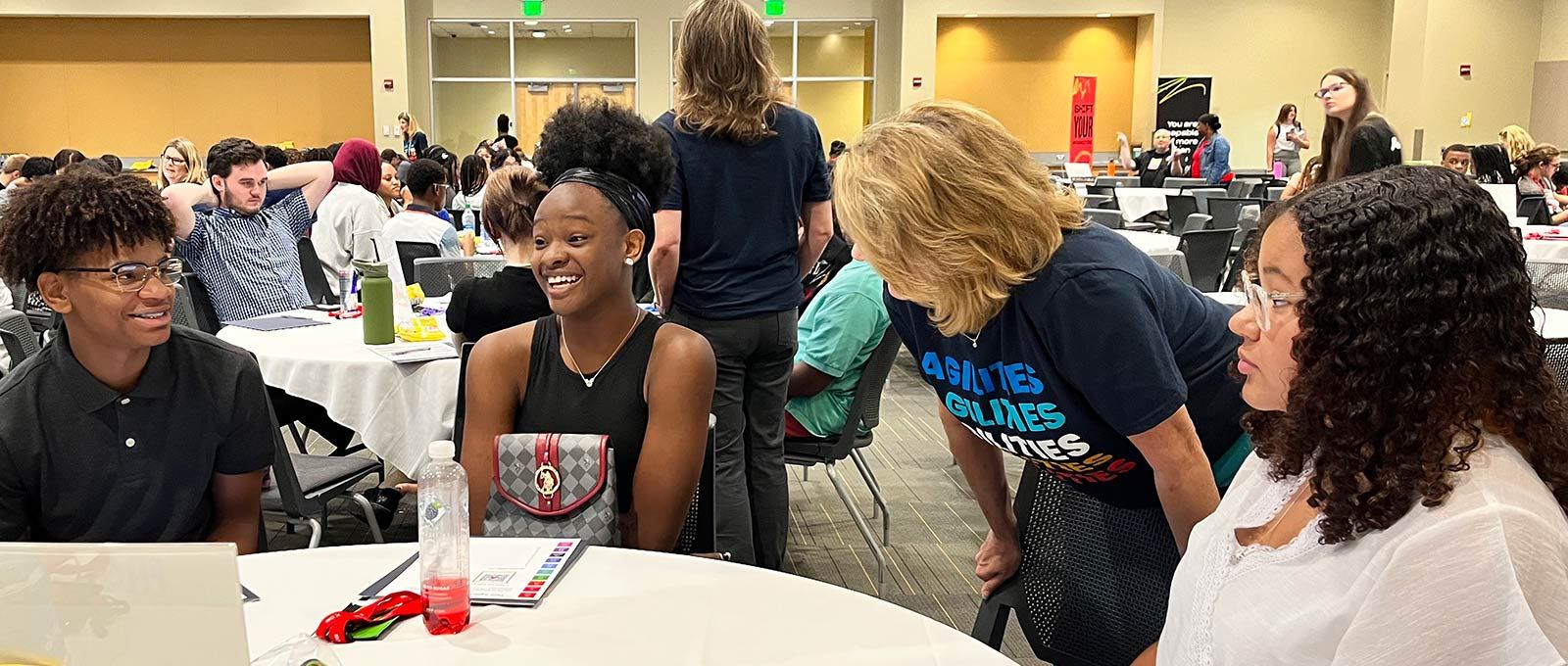 Three students with varying racial and ethnic backgrounds sit at a round table inside a conference room with hundreds of other peers.