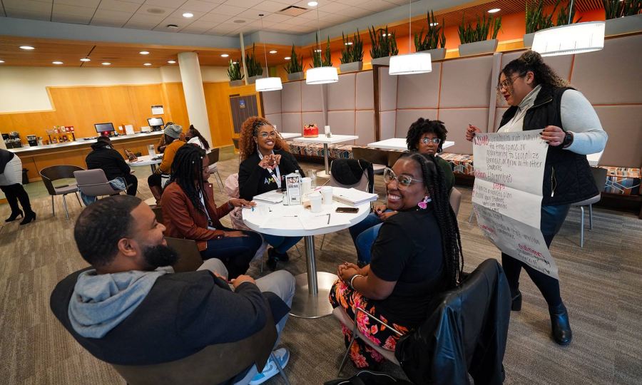 Five racially diverse Amplify attendees sit around a table while a sixth stands holding a large sheet of paper with notes written on it.